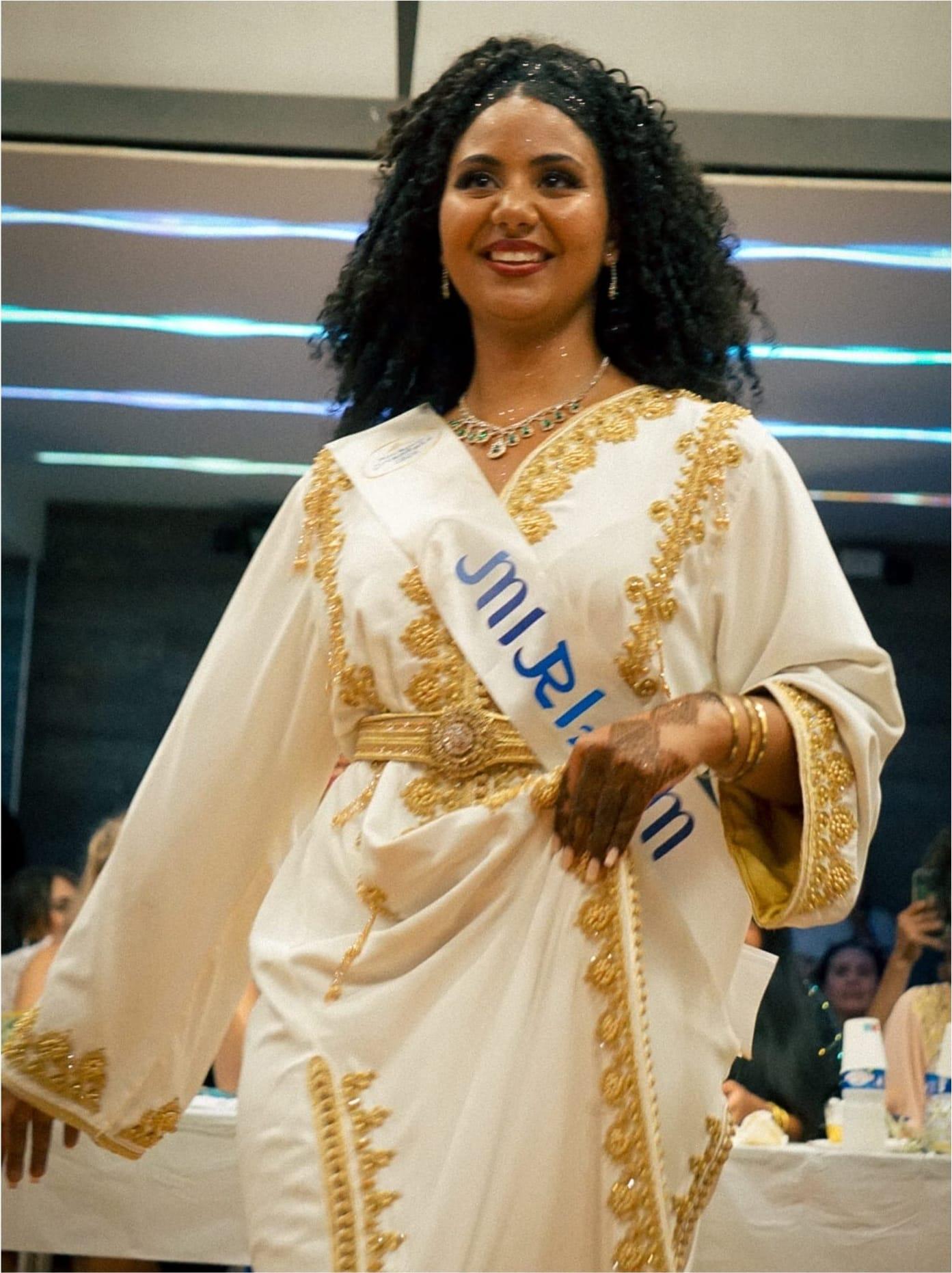 Une candidate souriante en caftan blanc brodé d’or et ceinture dorée, écharpe de miss en travers.