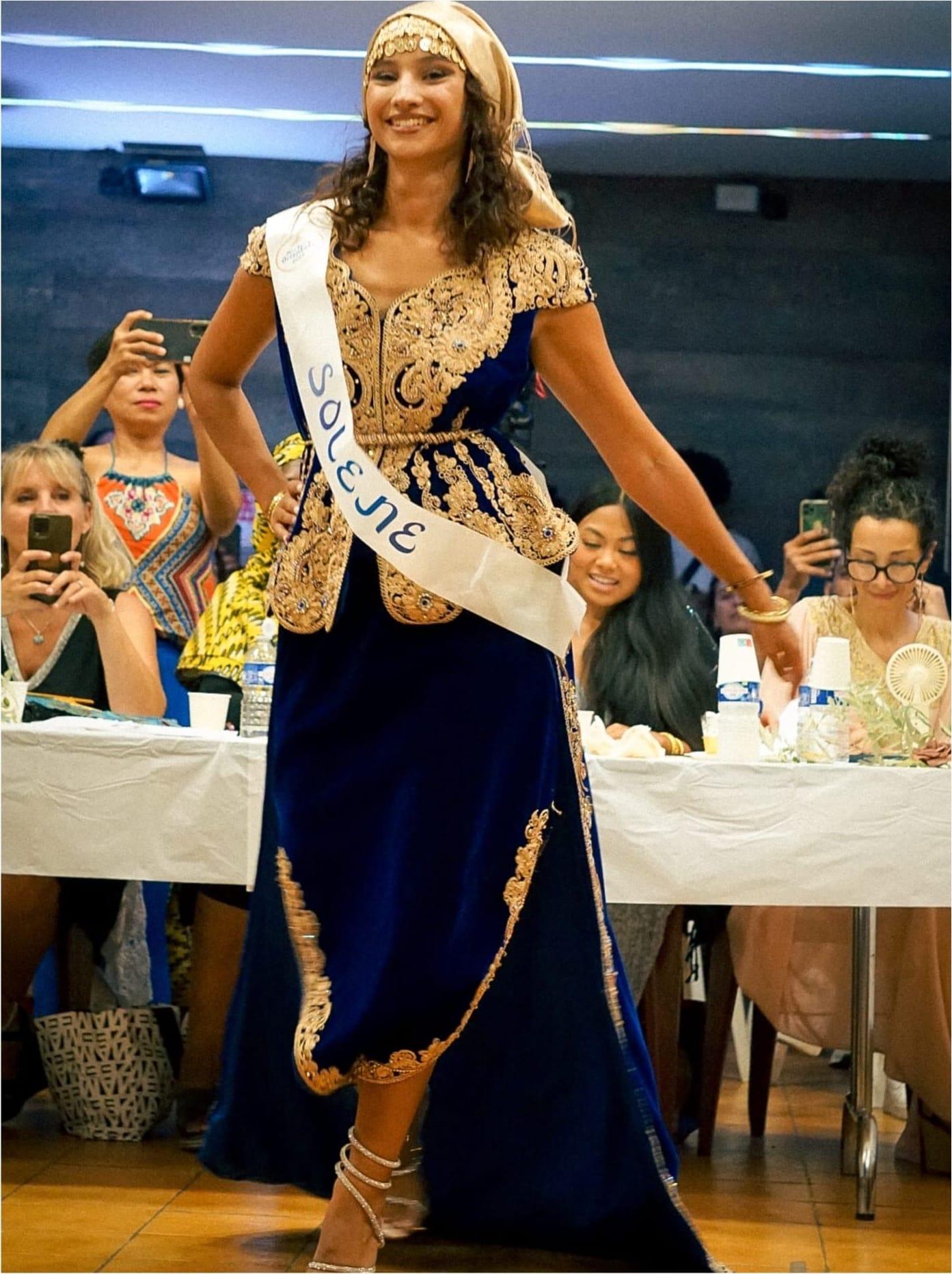 Une candidate en caftan bleu nuit et bustier brodé d’or, foulard doré dans les cheveux, défile entre les tables.