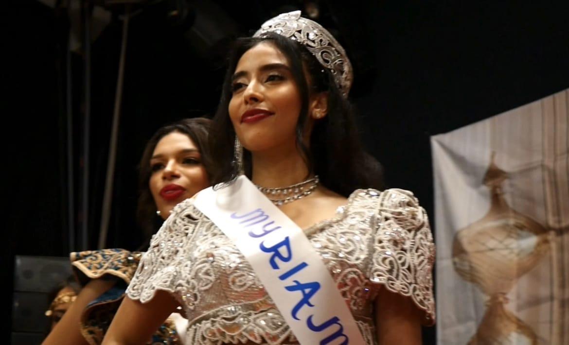 Une candidate coiffée d’un diadème, en robe blanche brodée et écharpe de miss, sourit sur scène.