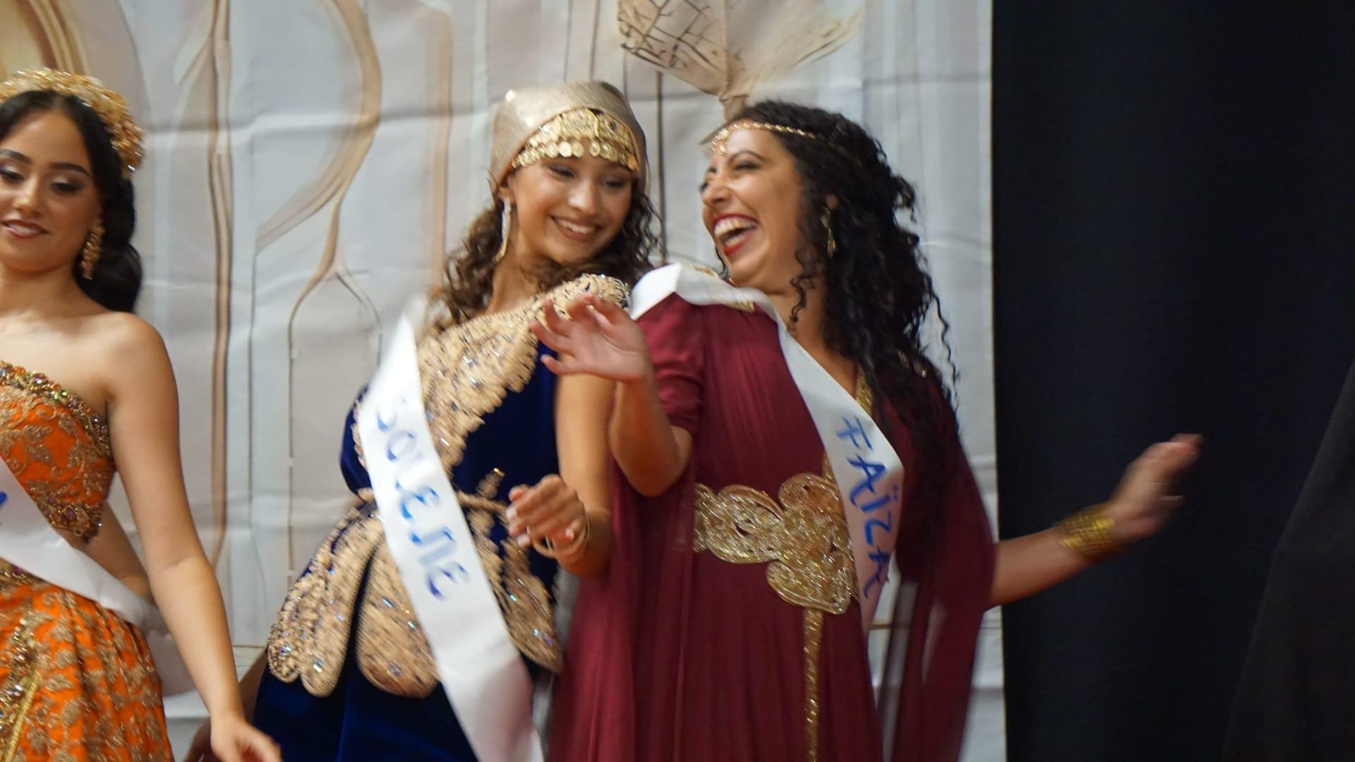 Des candidates en tenues traditionnelles et écharpes de miss rient ensemble sur scène.