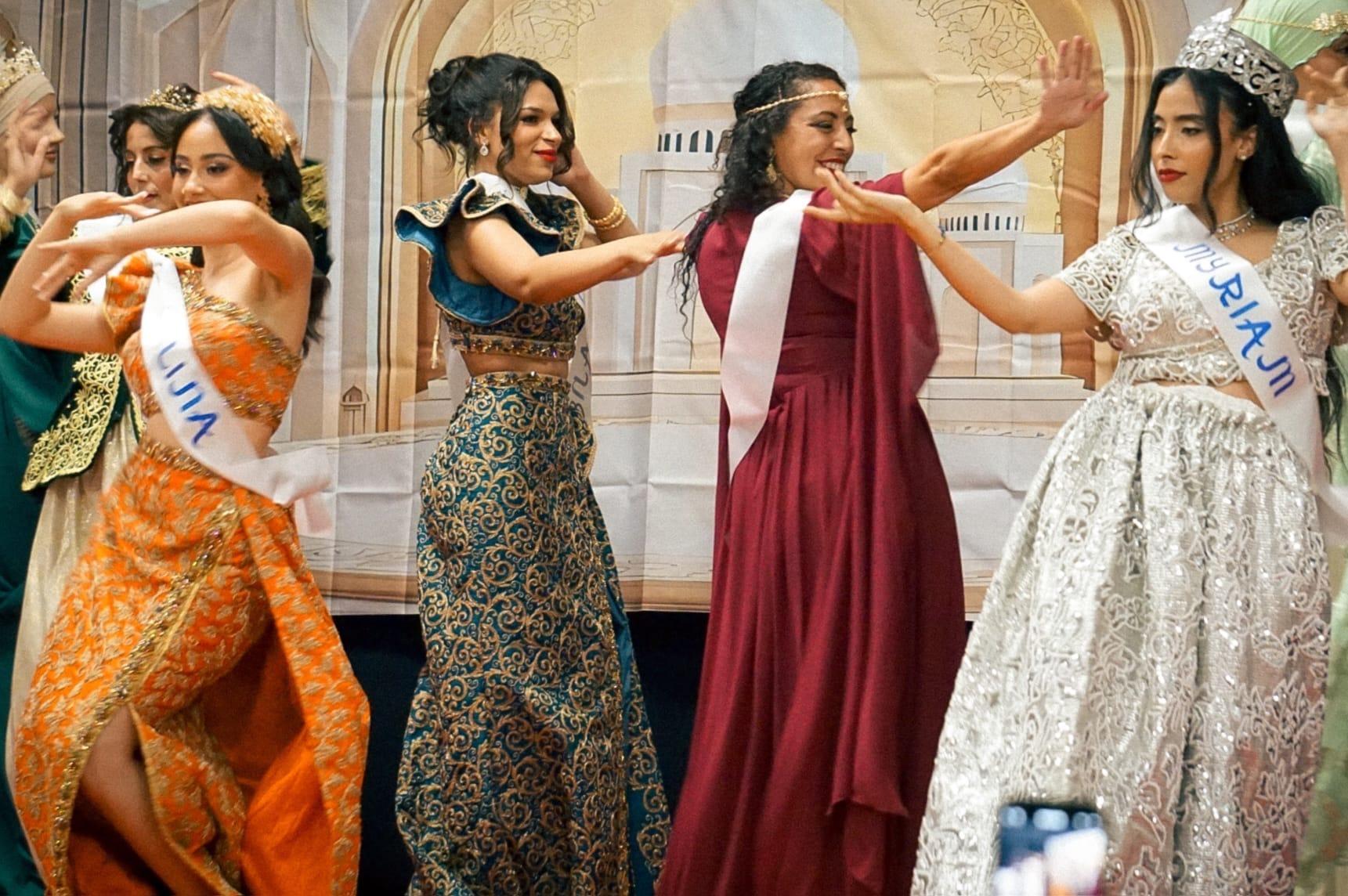 Danse des candidates sur scène : robes orange, bordeaux et argentée, sourires et mains en l’air.
