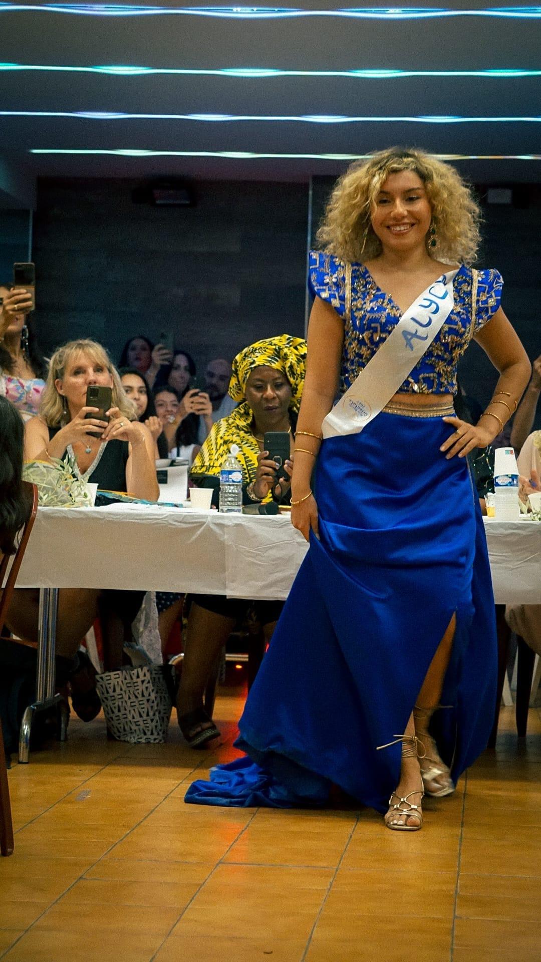 Une candidate aux cheveux bouclés, en tenue bleu roi brodée et écharpe de miss, pose devant les tables du public.