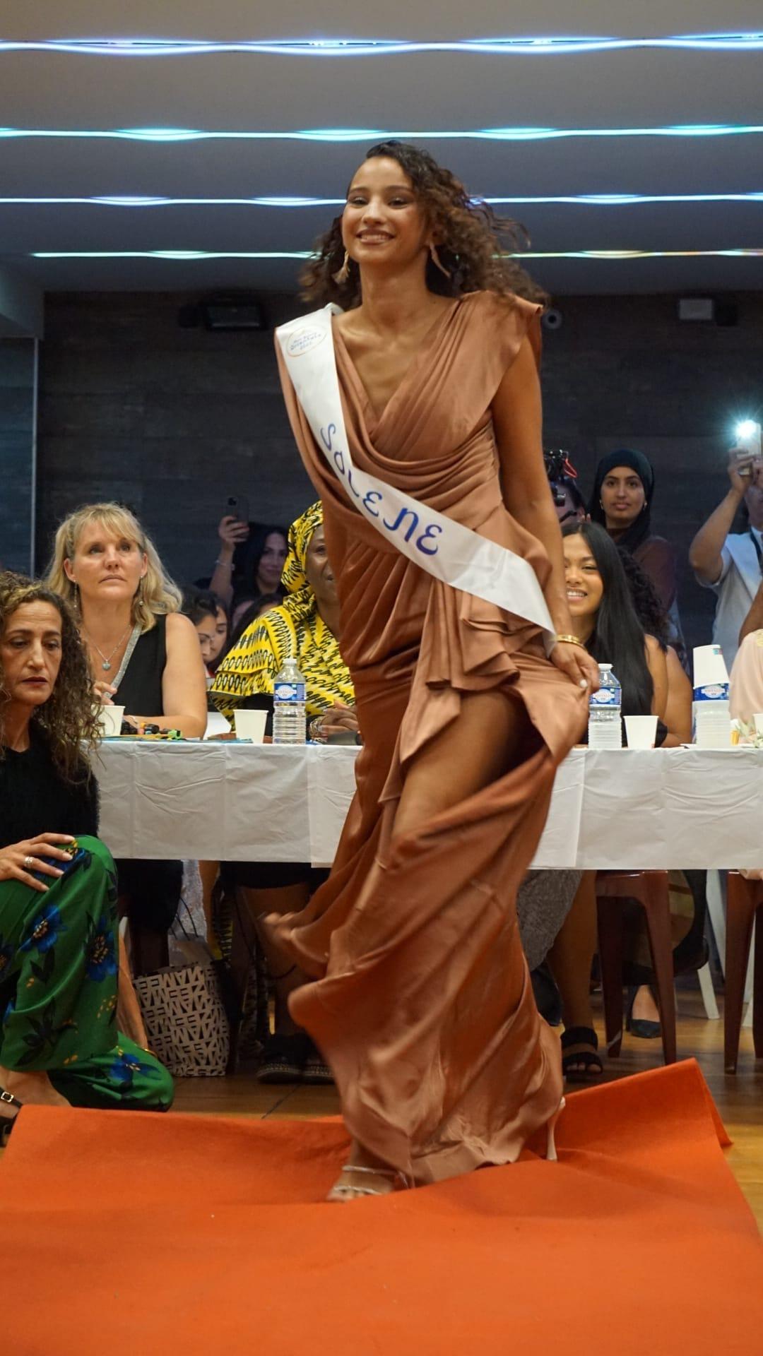 Une candidate en robe drapée couleur bronze et écharpe de miss défile sur le tapis orange, entre les tables du public.