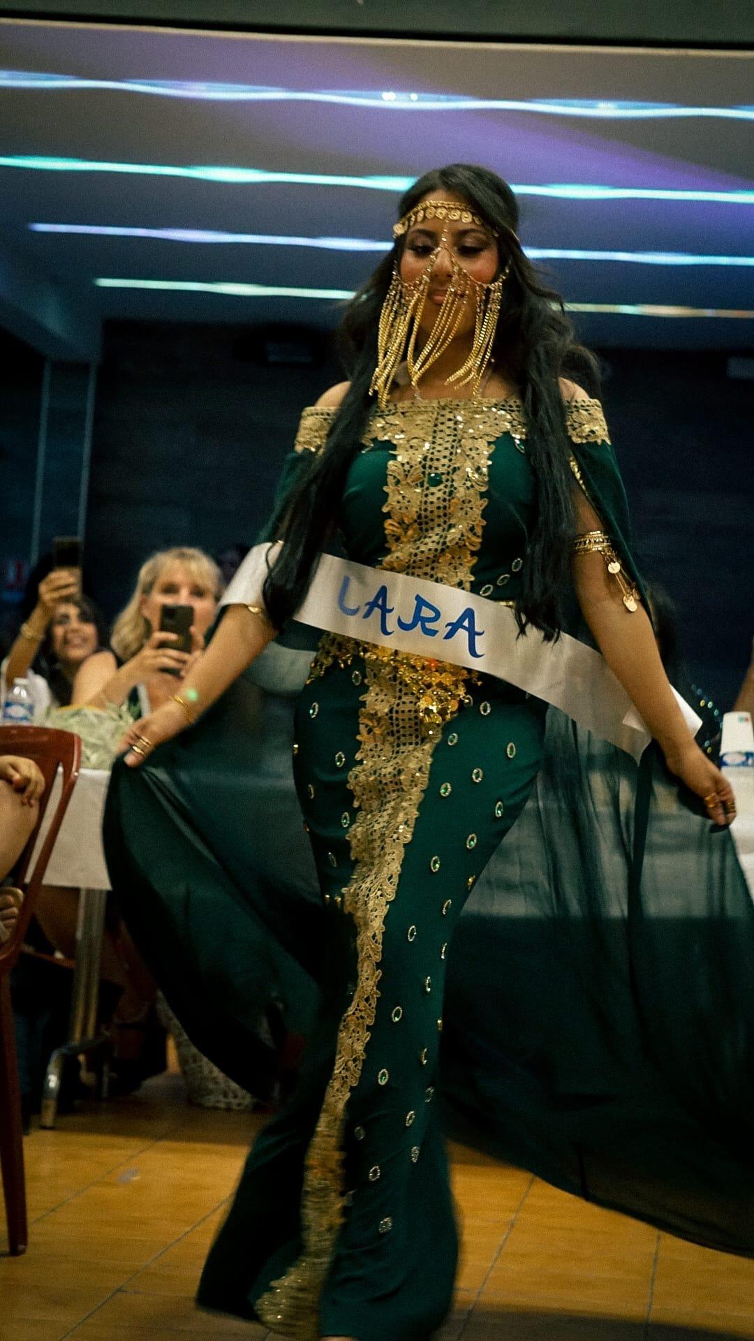 Une candidate voilée, en robe verte brodée d’or et écharpe de miss, défile devant le public qui la filme.