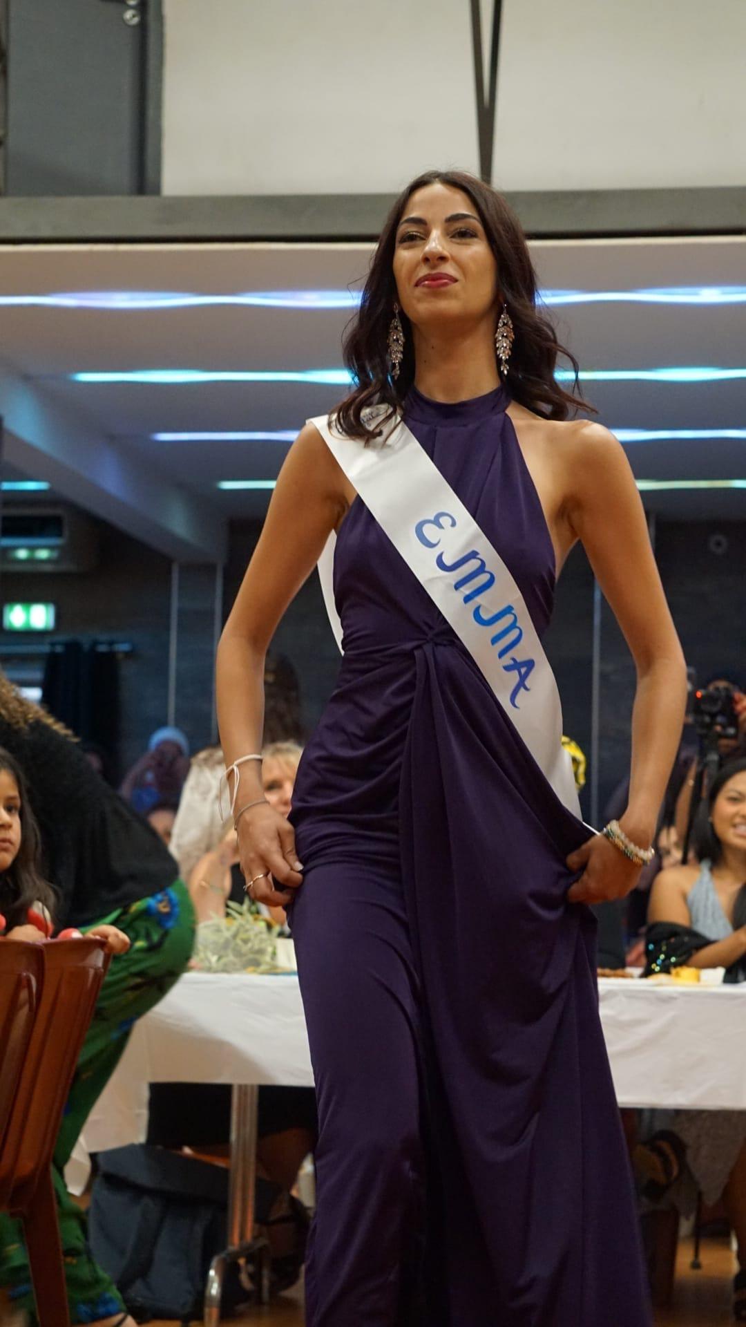 Une candidate en longue robe violette et écharpe de miss marche vers le public, cheveux lâchés.